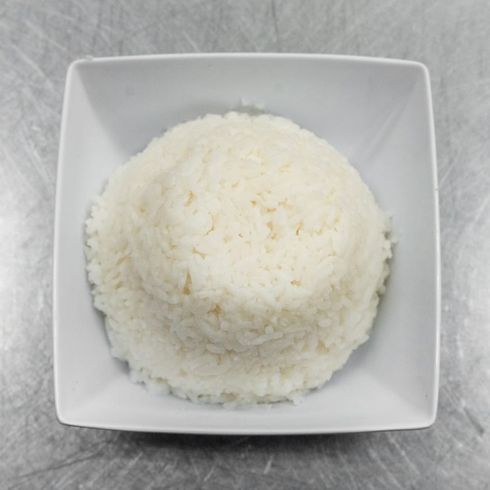 A white square bowl filled with fluffy white rice, centered on a metallic surface.