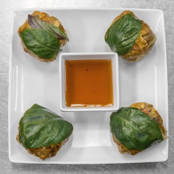 Four basil-wrapped dumplings with a small square dish of dipping sauce on a white plate.