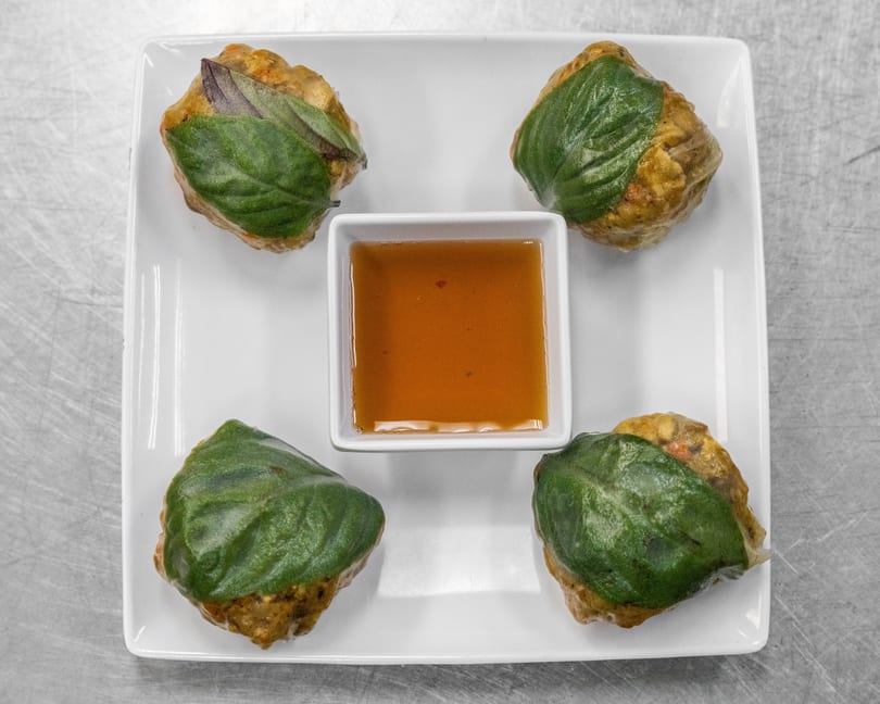 Four basil-wrapped dumplings with a small square dish of dipping sauce on a white plate.