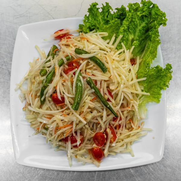 A white plate of Som Tum (Thai papaya salad) with green beans, tomatoes, and lettuce.