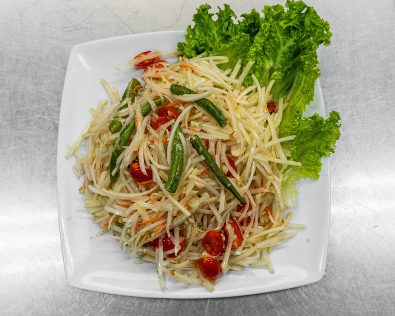 A white plate of Som Tum (Thai papaya salad) with green beans, tomatoes, and lettuce.