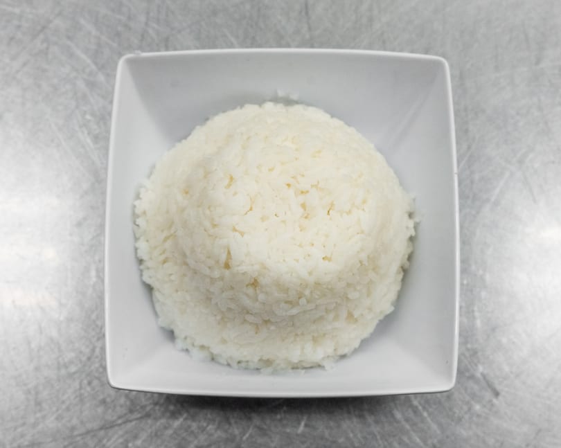 A white square bowl filled with fluffy white rice, centered on a metallic surface.