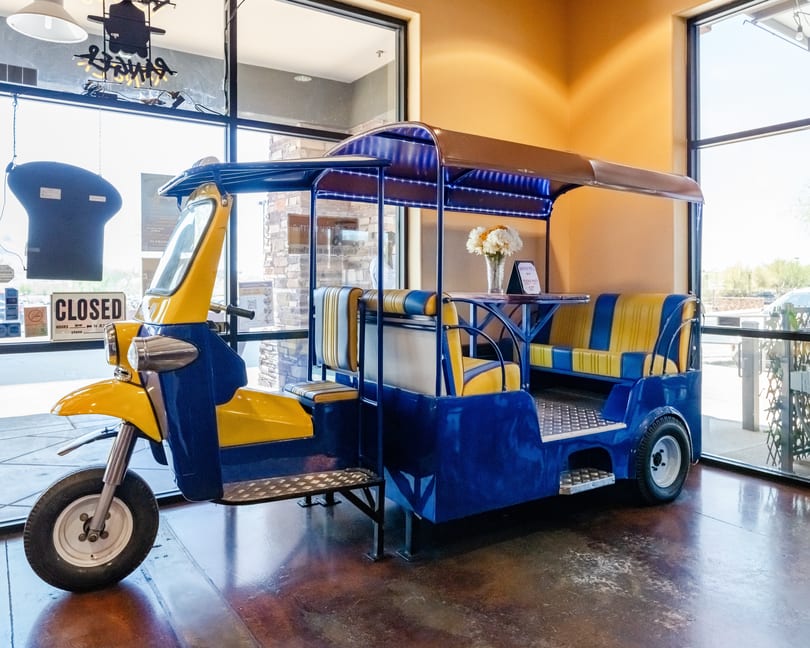 A blue and yellow tuk-tuk with seating and a small table, parked indoors near a window.