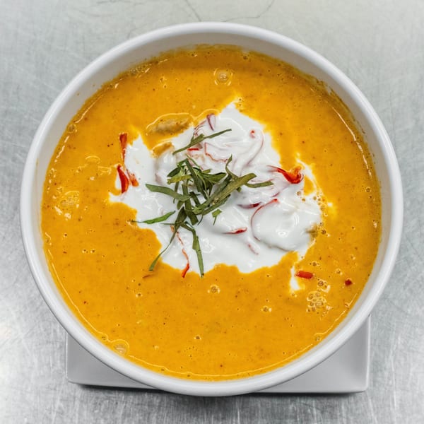 A white bowl of orange curry with white cream, red peppers, and green herbs on top.