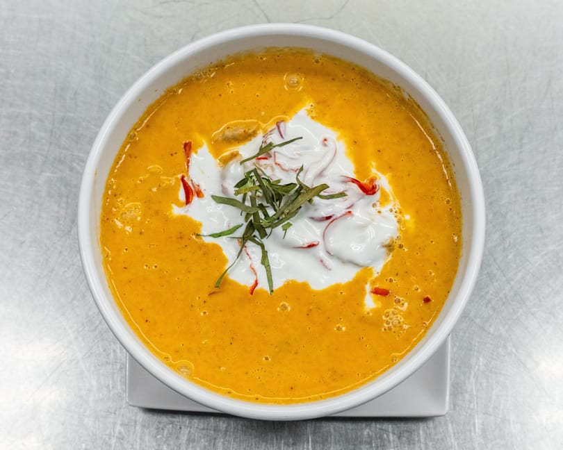 A white bowl of orange curry with white cream, red peppers, and green herbs on top.