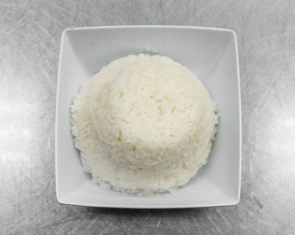A white square bowl filled with a mound of fluffy white rice on a metallic surface.