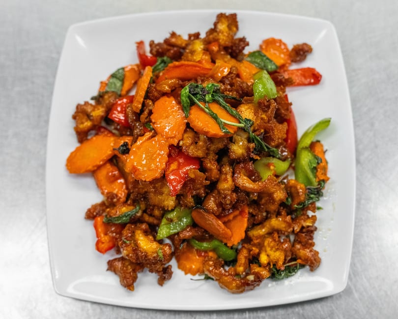 Crispy chicken stir-fry with carrots, bell peppers, and basil on a white rectangular plate.